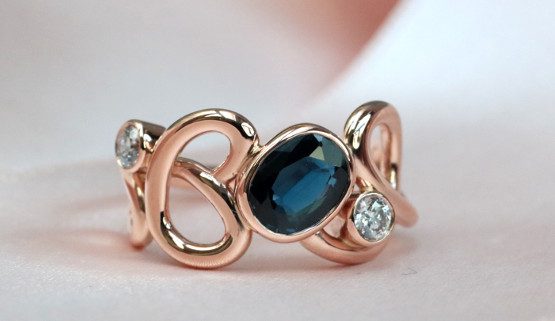 verlobungsring aussergewöhnlich Ein Rotgoldfaden schlingt sich um den Finger, in den ein ovaler Saphir und zwei Diamanten eingearbeitet sind.