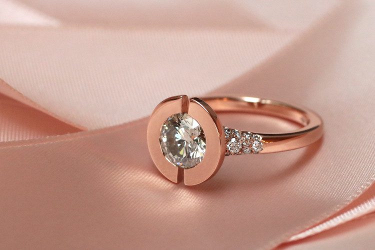 Un disque en or rose est scindé en deux, avec une ouverture verticale au milieu. Un diamant est serti dans ce disque. La bague montre un style de créateur moderne et contemporain. Un pavage de diamant orne le corps de la bague créee sur-mesure des deux côtés.