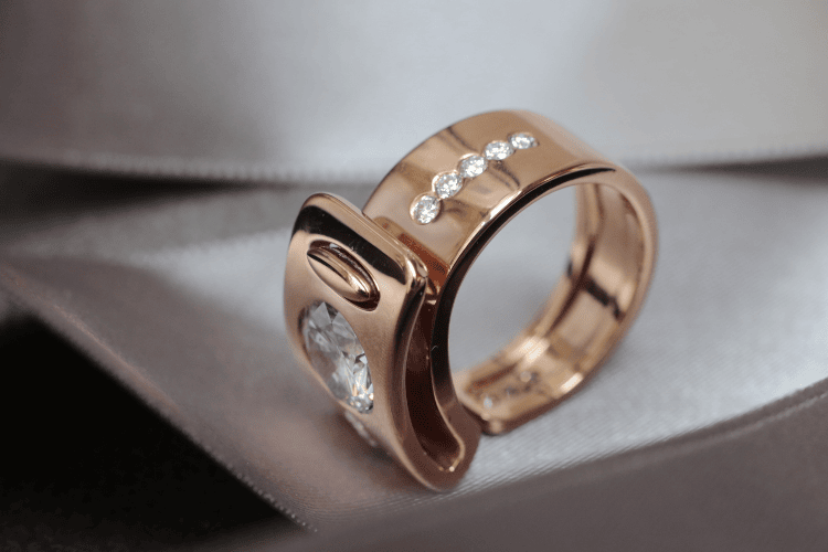 Un ruban en or rouge forme un cinq, vu de profile. Cette bague pour femme affrirmée est sertie de diamants.