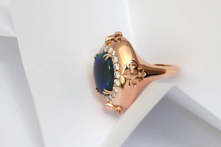 Il s'agit d'une bague sur-mesure opulante, ovale, sertie d'une opale noire d'Australie, revelant des reflèts verts sur fond bleu, telle une vague de fond de l'Océan. Autour de l'opale, des diamants, disposés en cercle la mettent encore plus en valeur. Quatre fleurs de lys sculptés dans de l'or rouge l'enjolivent sur les côtés. C'est une création de bague sur-mesure de la joaillière Annette Girardon, à Paris.
