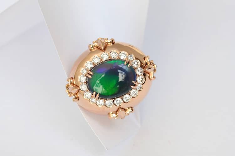 Cette bague ovale, d'un volume généreux, est sertie d'une opale aux reflèts verts sur fond bleu, rappelant une vague de fond de l'Océan. Des diamants sont serties en cercle autour de l'opale. Quatre fleurs de lys en or rouge complètent ce design, inventé par la joaillière Annette Girardon, installée à Paris.