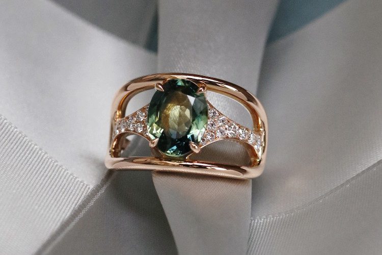 Un saphir bi-colore ovale orne le centre de cette bague créée sur-mesure. Deux éléments pavés de diamants le relie à une élipse en fil d'or rouge.